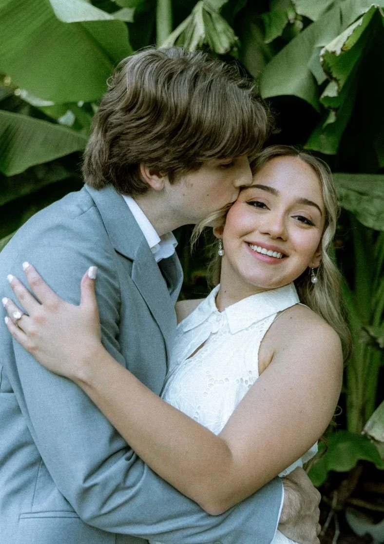 A young couple embracing outdoors, with the man kissing the woman's cheek and the woman smiling happily. Behind them are large green tropical leaves.