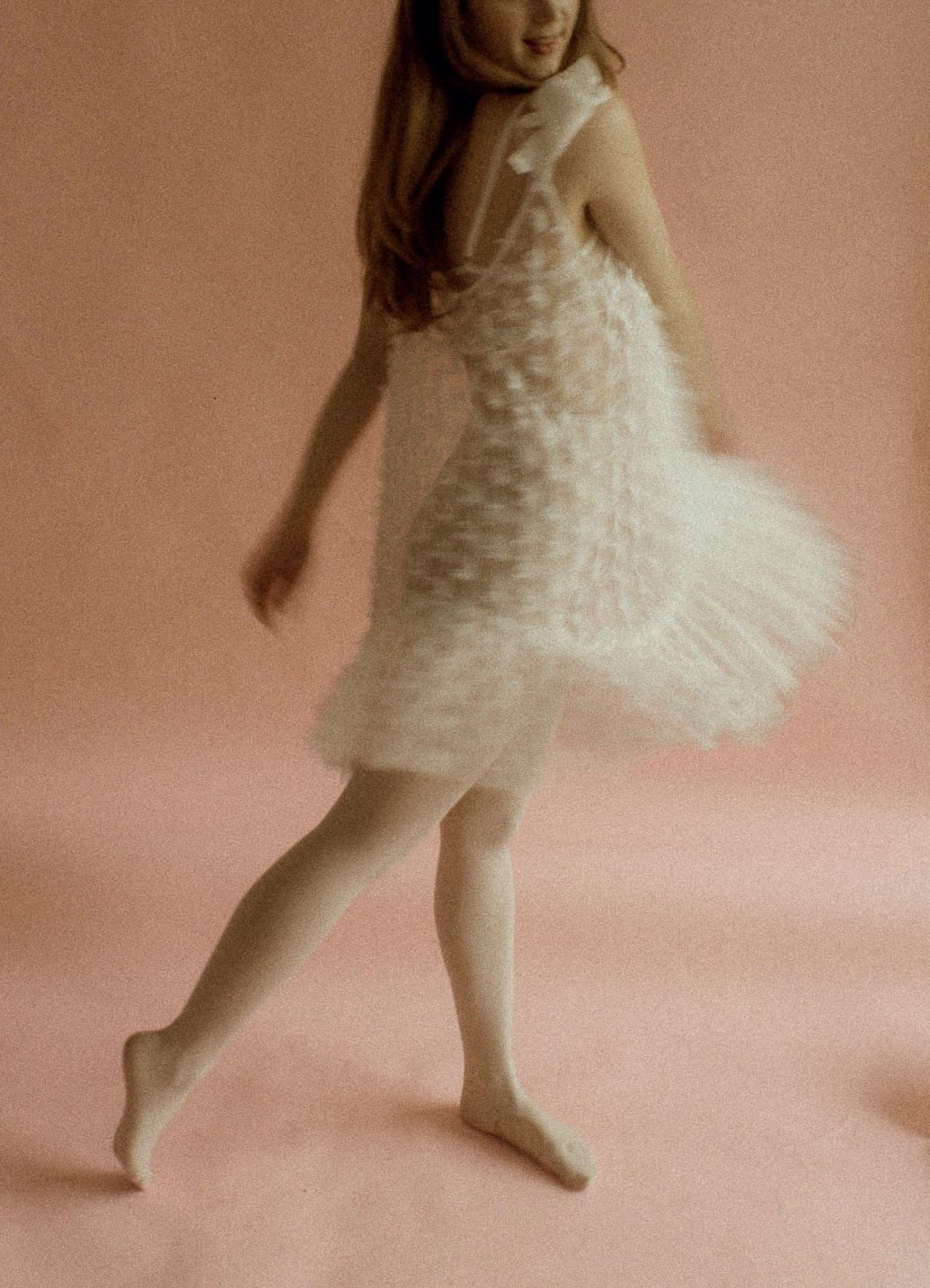 A woman in a white fluffy dress and beige tights standing on a light pink background, with her head slightly turned and her right hand touching her thigh.