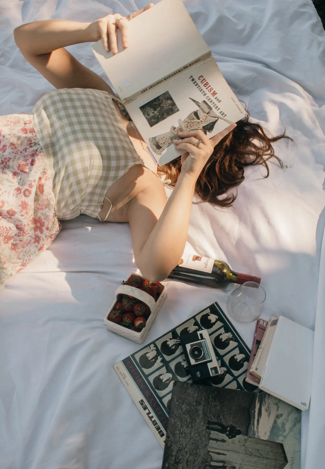 A woman lying on a bed reading a book, with a bottle of wine, a glass, strawberries, books, a camera, and a box of matches nearby.
