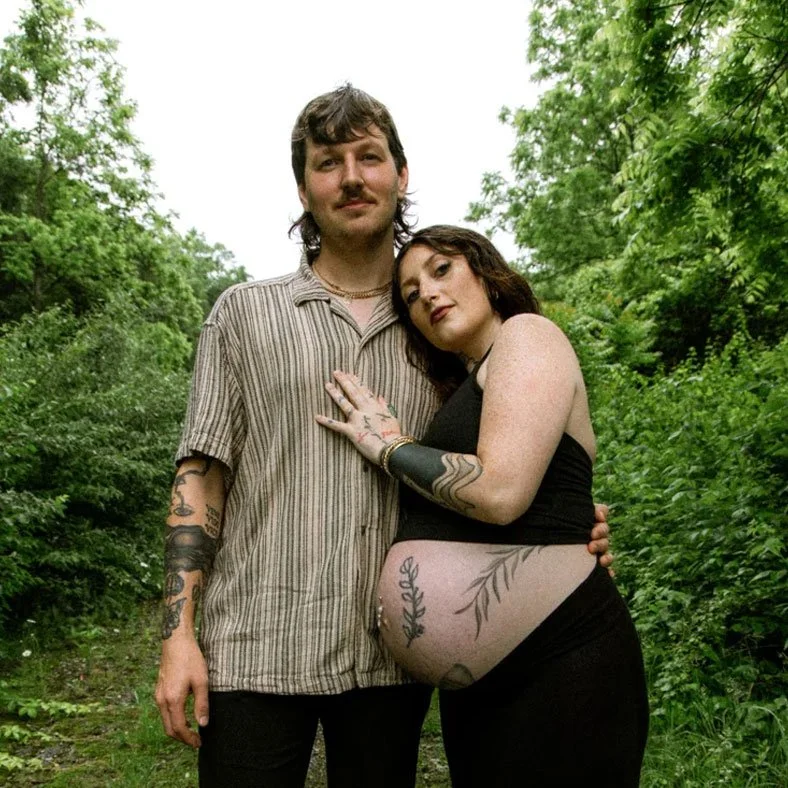 A couple standing together outdoors in a lush green wooded area, smiling at the camera. The woman, who is pregnant, has tattoos on her arm and belly, and the man has tattoos on his arm.