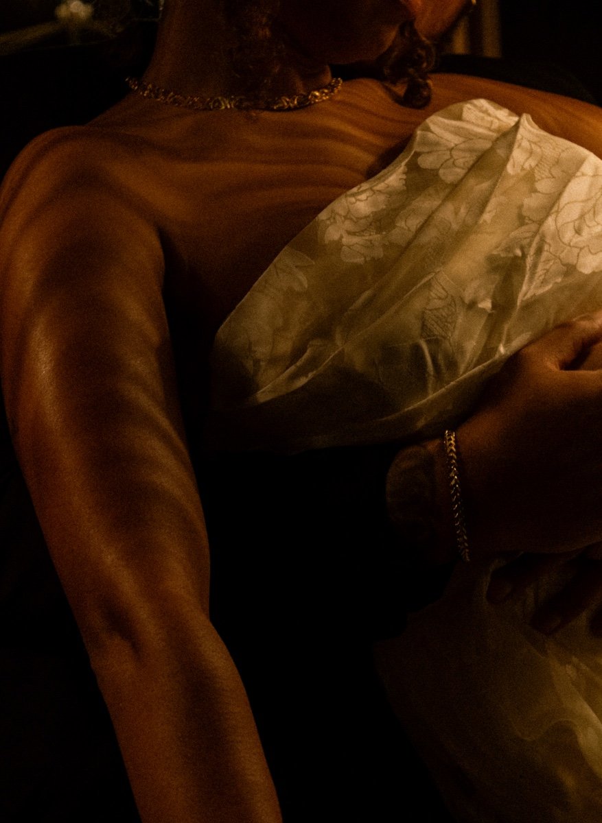 A woman in a shiny, gold-colored dress with a floral scarf around her neck, sitting in a dimly lit environment, holding her phone.