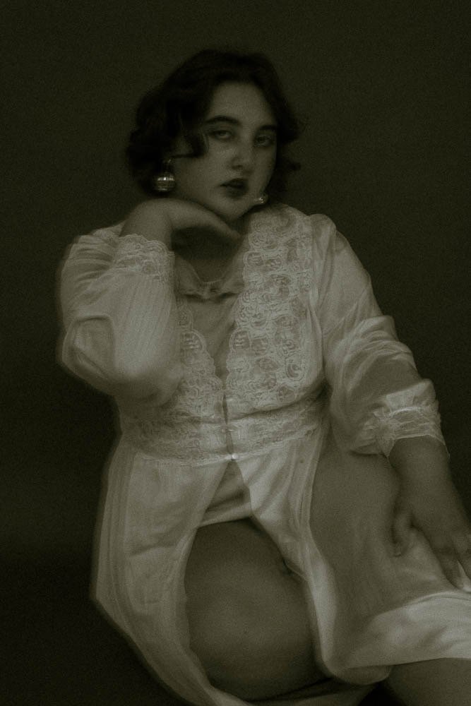 A woman wearing vintage-style lace blouse and earrings, sitting with one hand resting near her face against a dark background.