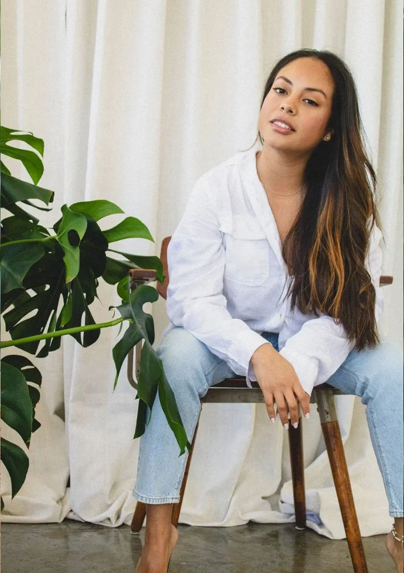 A woman with long brown hair sits on a wooden chair in front of a cream-colored curtain, wearing a white shirt and light blue jeans, sitting with one leg crossed and leaning slightly to the side. A large green plant is partially visible on her left.