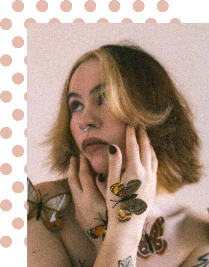 A woman with short, wavy hair and multiple tattoos, including a butterfly tattoo on her chest, is surrounded by live butterflies landing on her hands and shoulders. She has dark lipstick, a septum piercing, and is gazing upward with a thoughtful expression.
