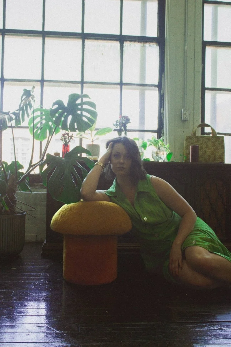 A woman in a green dress resting on a mushroom-shaped cushion in front of large window and green plants.