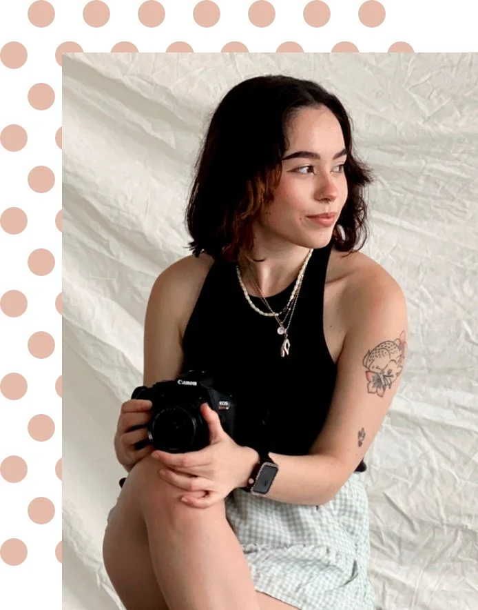 Young woman with dark hair and tattoos wearing a black top, pearl necklace, and smartwatch, holding a Canon camera, sitting against a crinkled white background.