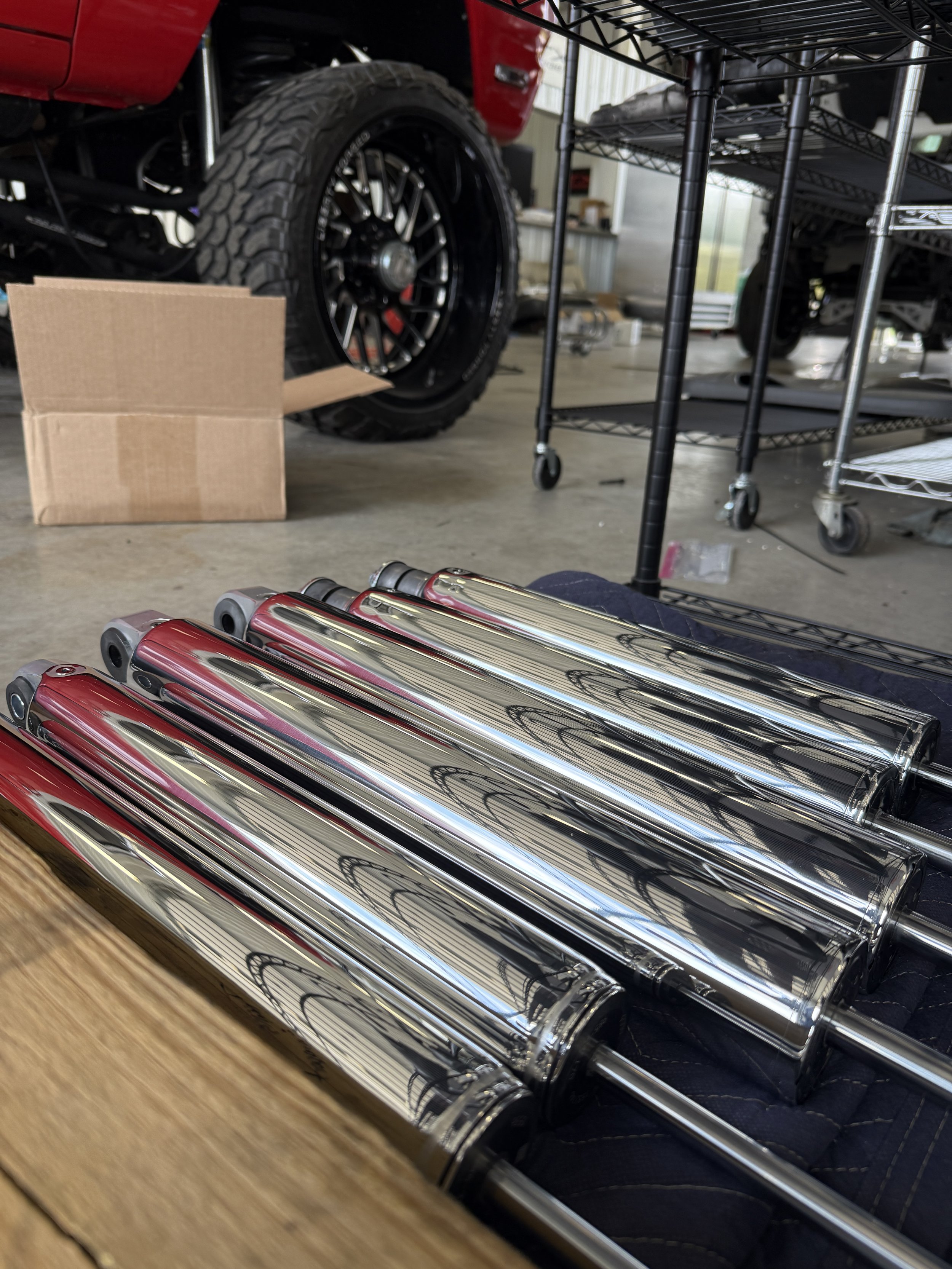 Close-up of shiny chrome motorcycle exhaust pipes on a wooden surface with a metal shelving unit and black tires in the background.