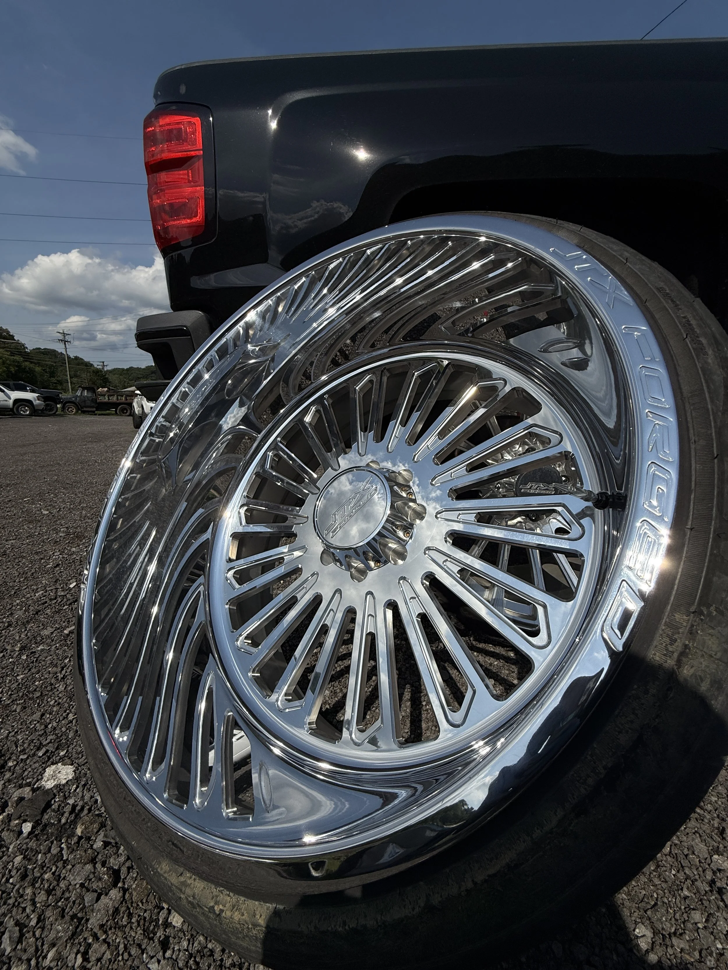 Close-up of chrome car rims with black tires on a black truck, outdoors on a sunny day with a clear blue sky and white clouds.