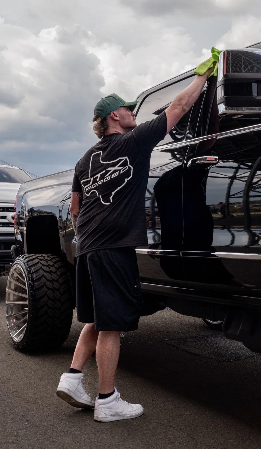 A man cleaning the window of a black pickup truck with a yellow cloth at an outdoor parking lot, with gray clouds overhead.