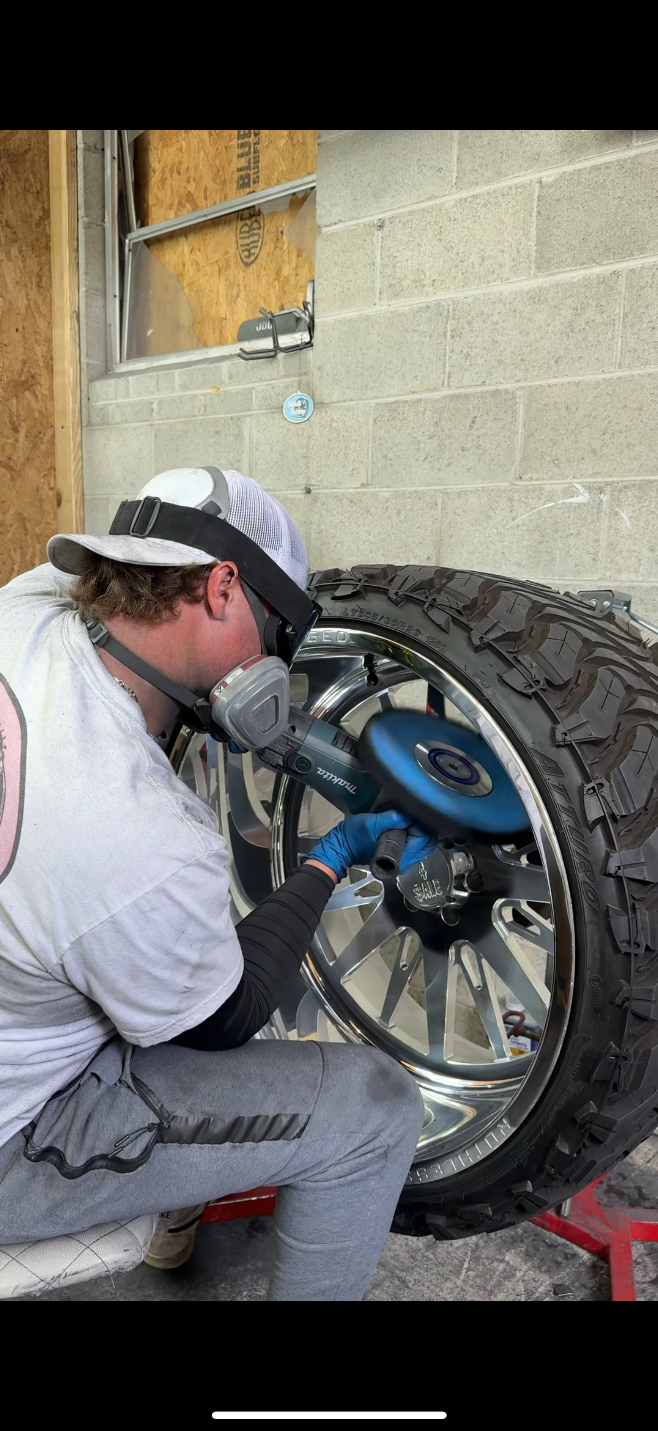 A person working on polishing a large, shiny car wheel with an electric polisher in a garage or workshop.