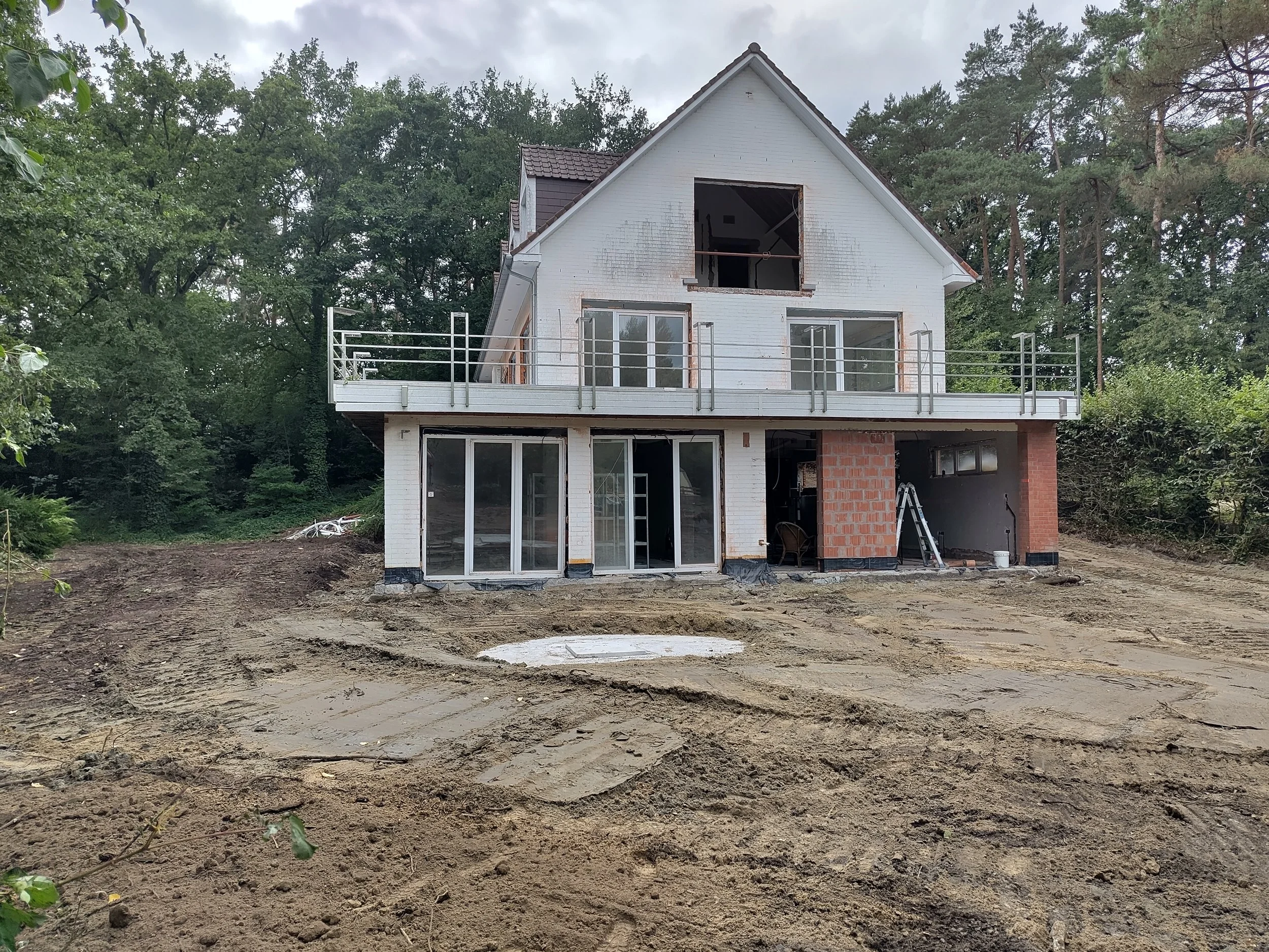 A house under construction with white brick walls, glass sliding doors, and an unfinished upper story with a large open window. The area around the house has bare dirt and construction materials, with trees in the background.