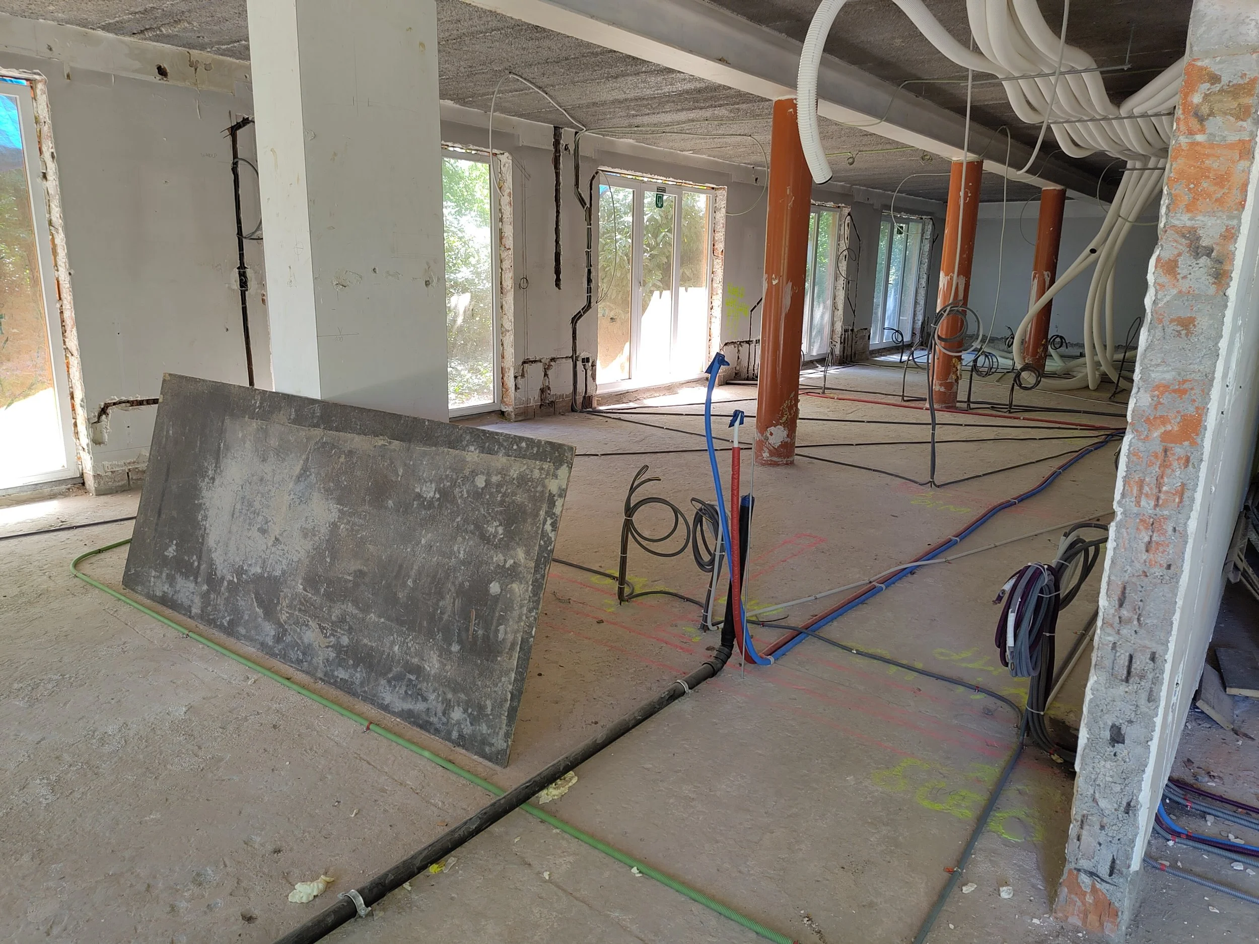 Interior of a building under construction with exposed pipes, electrical wiring, large windows, and unfinished walls.