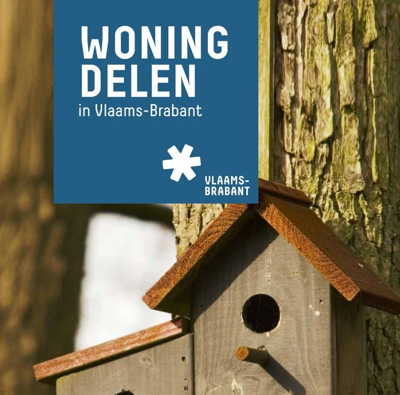 A blue sign with white text reading "Woning Delen in Vlaams-Brabant" and a white star icon, attached next to a wooden birdhouse mounted on a tree trunk.
