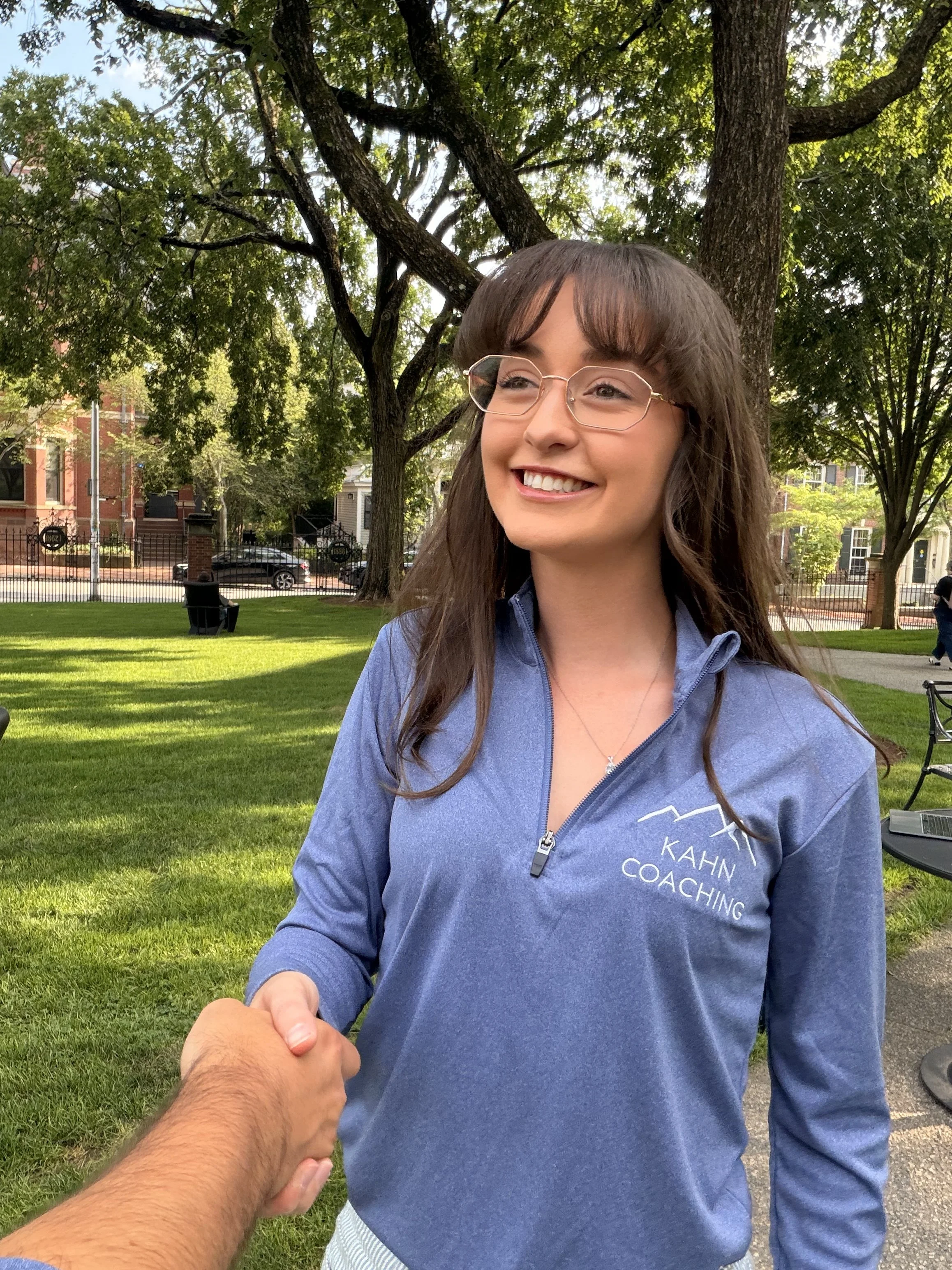 A young woman with long brown hair, glasses, and a blue jacket smiling and holding hands with someone outside in a park with green grass, trees, buildings, and cars in the background.
