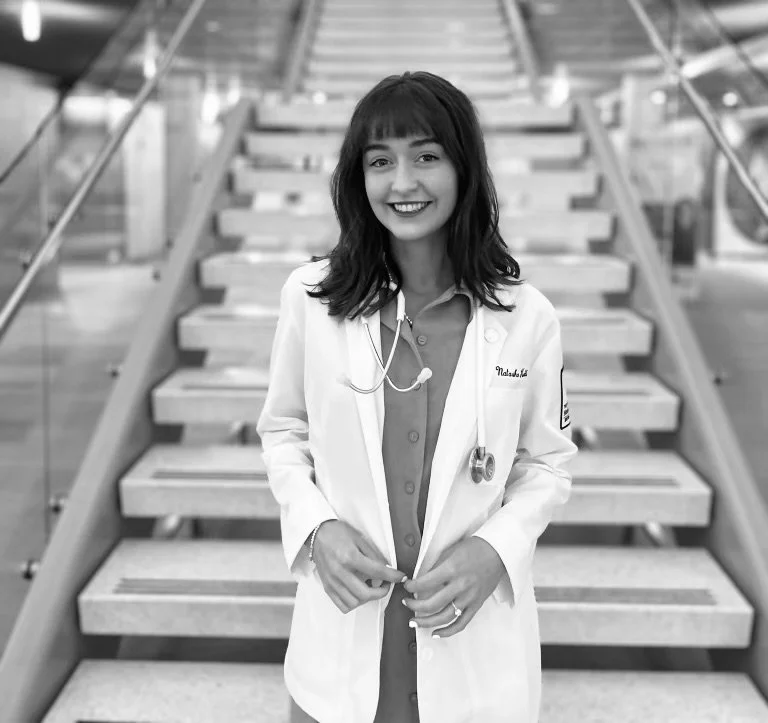 A smiling female doctor or nurse in a white coat standing on a staircase in a medical building.