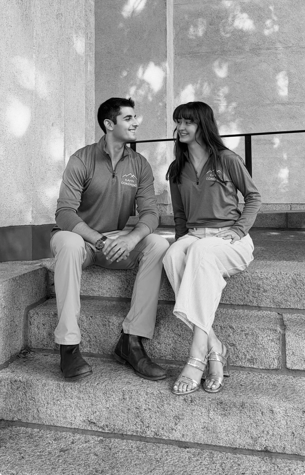A black-and-white photo of a young man and woman sitting on stairs outside, facing each other, smiling. Both are wearing matching zip-up jackets with a mountain logo and the words 'KAHN COACHING.' The man is wearing light-colored pants and boots, while the woman is wearing a skirt and sandals.