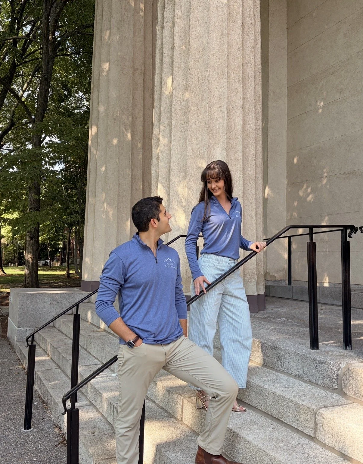 Two young adults, a man and a woman, standing and sitting on steps outside a building with columns, engaged in conversation and smiling at each other, wearing matching blue shirts.