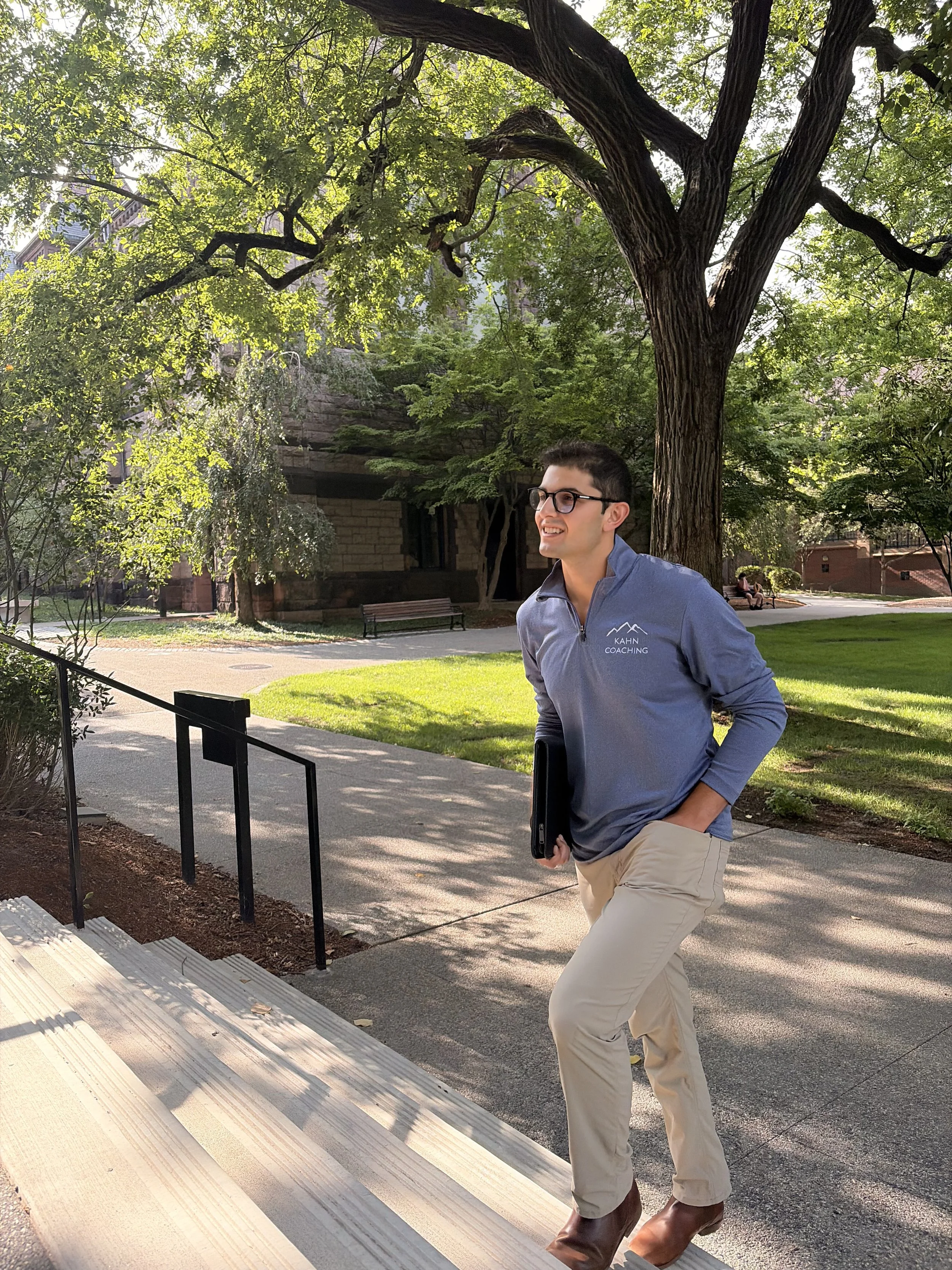 A man wearing glasses, a gray jacket with 'KAHN COACHING' logo, beige pants, and brown boots standing near stairs outside in a park or campus area, with green trees and a bench in the background.