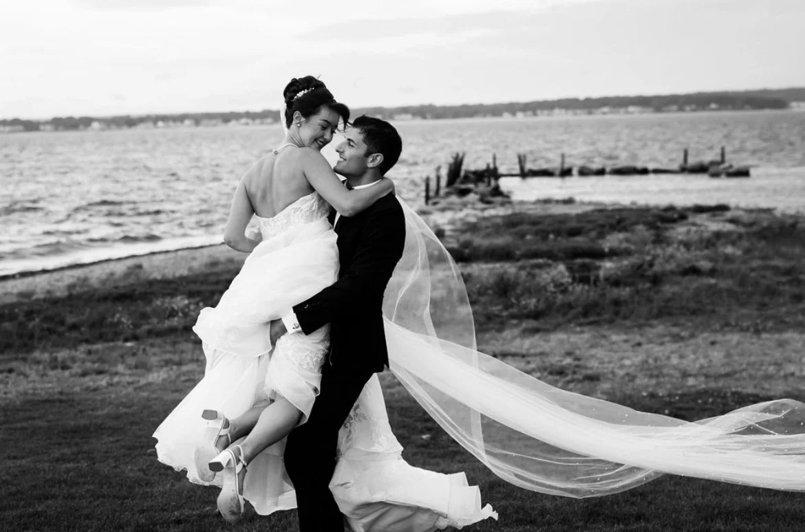 A black and white photo of a bride and groom celebrating outdoors by the water. The groom is lifting the bride, who is smiling and hugging him, with a blurred pier in the background.