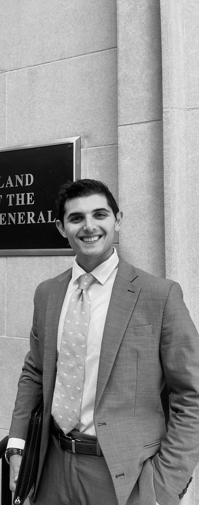 Young man in a suit and tie smiling standing outside a government building with a sign on the wall.