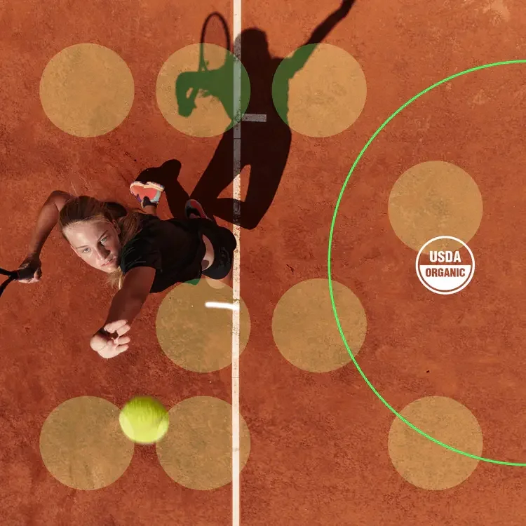 A female tennis player on a clay court trying to hit a yellow tennis ball with a racket, with her shadow visible on the court and a USDA Organic label.