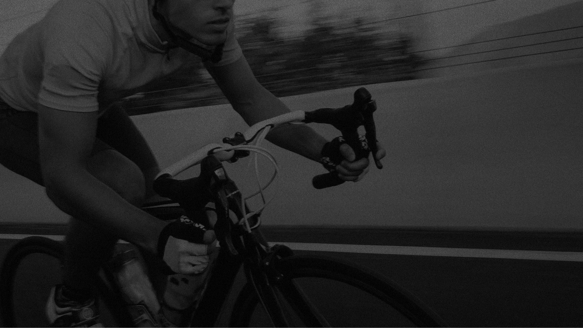 Black and white photo of a person riding a bicycle, holding a bottle in their right hand, with a chain-link fence and trees in the background.