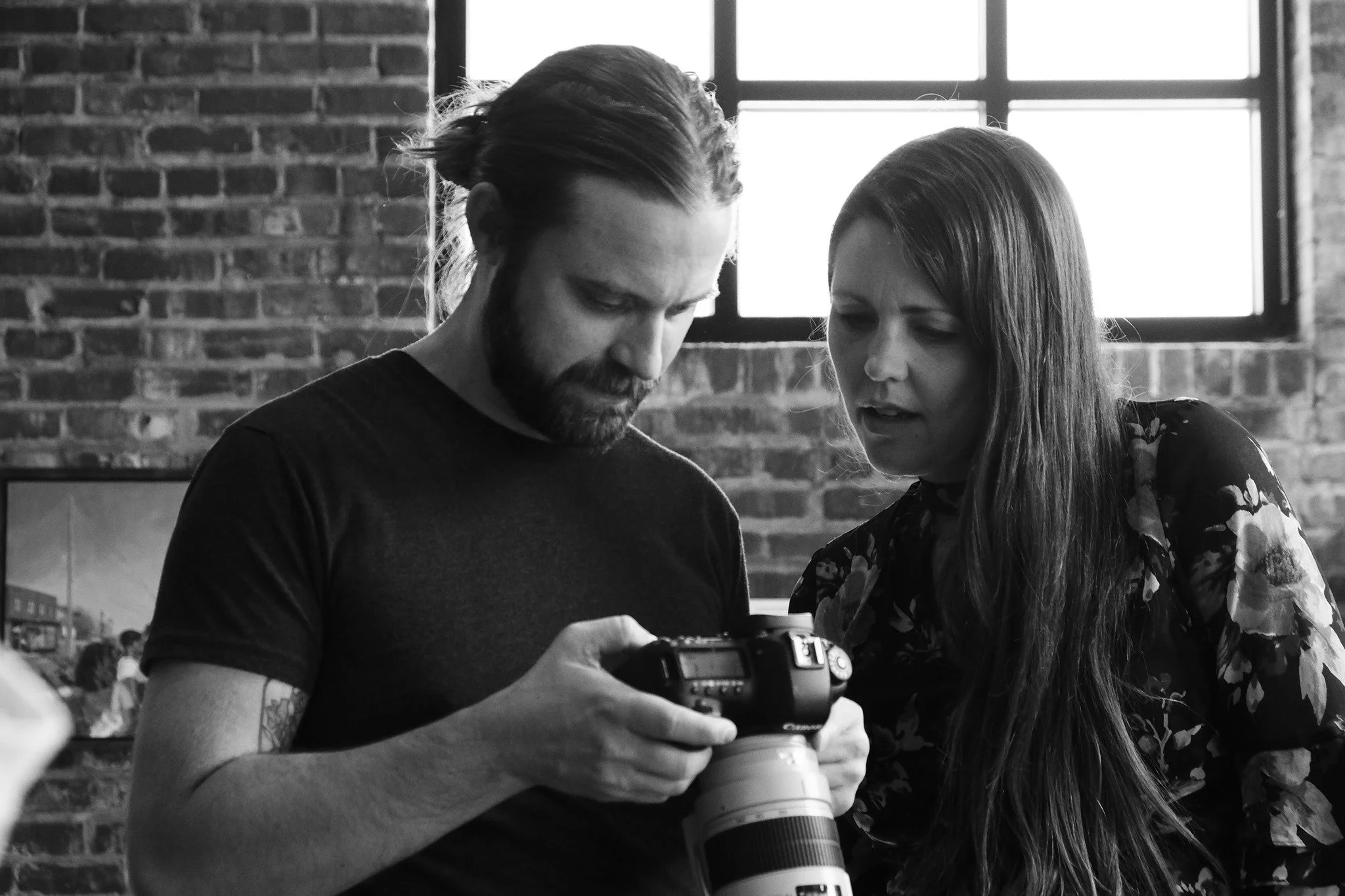 A man and woman looking at a camera, with a brick wall and window in the background.