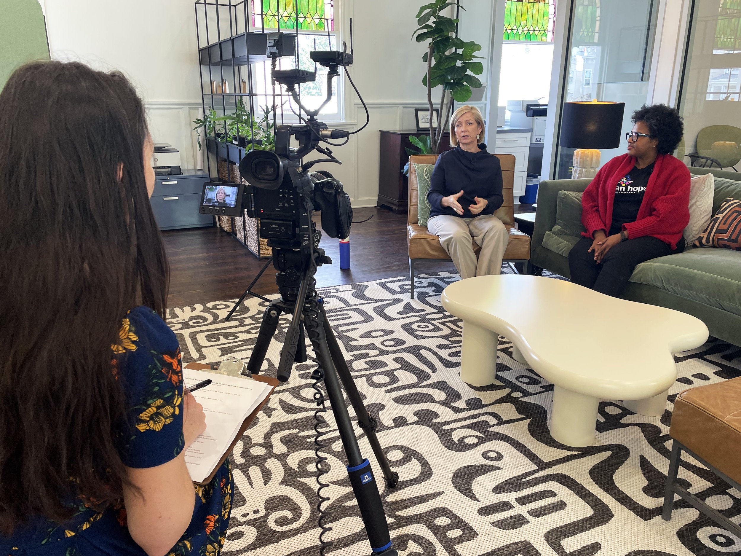 Two women seated and talking during an interview or conversation, with a camera mounted on a tripod recording them. One woman has blonde hair and is wearing a dark sweater, while the other has curly hair, glasses, and is wearing a red cardigan over a black shirt with a colorful logo. They are in a brightly lit room with plants, furniture, and large windows in the background.