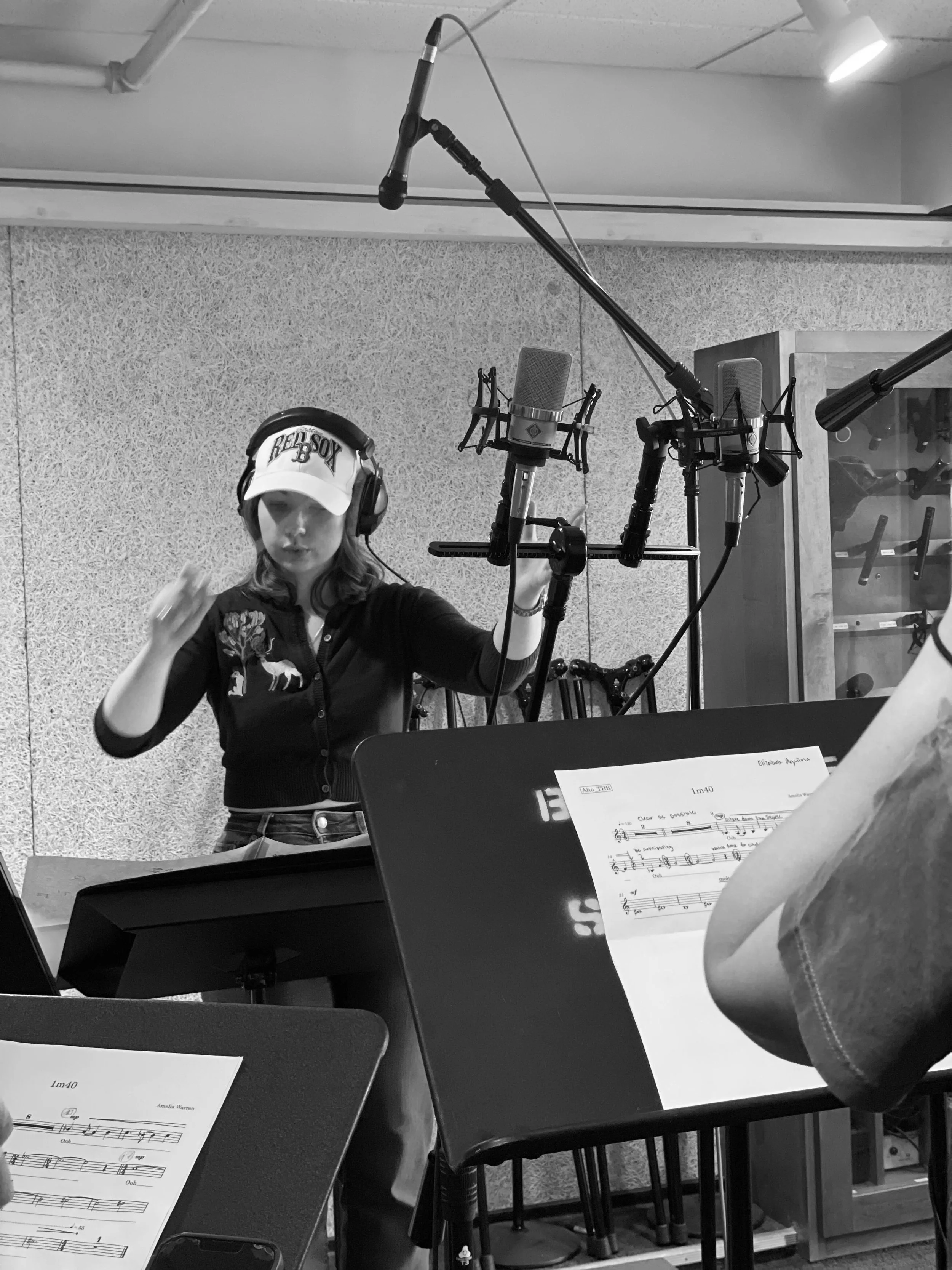 A woman in a recording studio wearing headphones and a cap, standing behind a microphone, with sheet music on a music stand and musical instruments in the background.