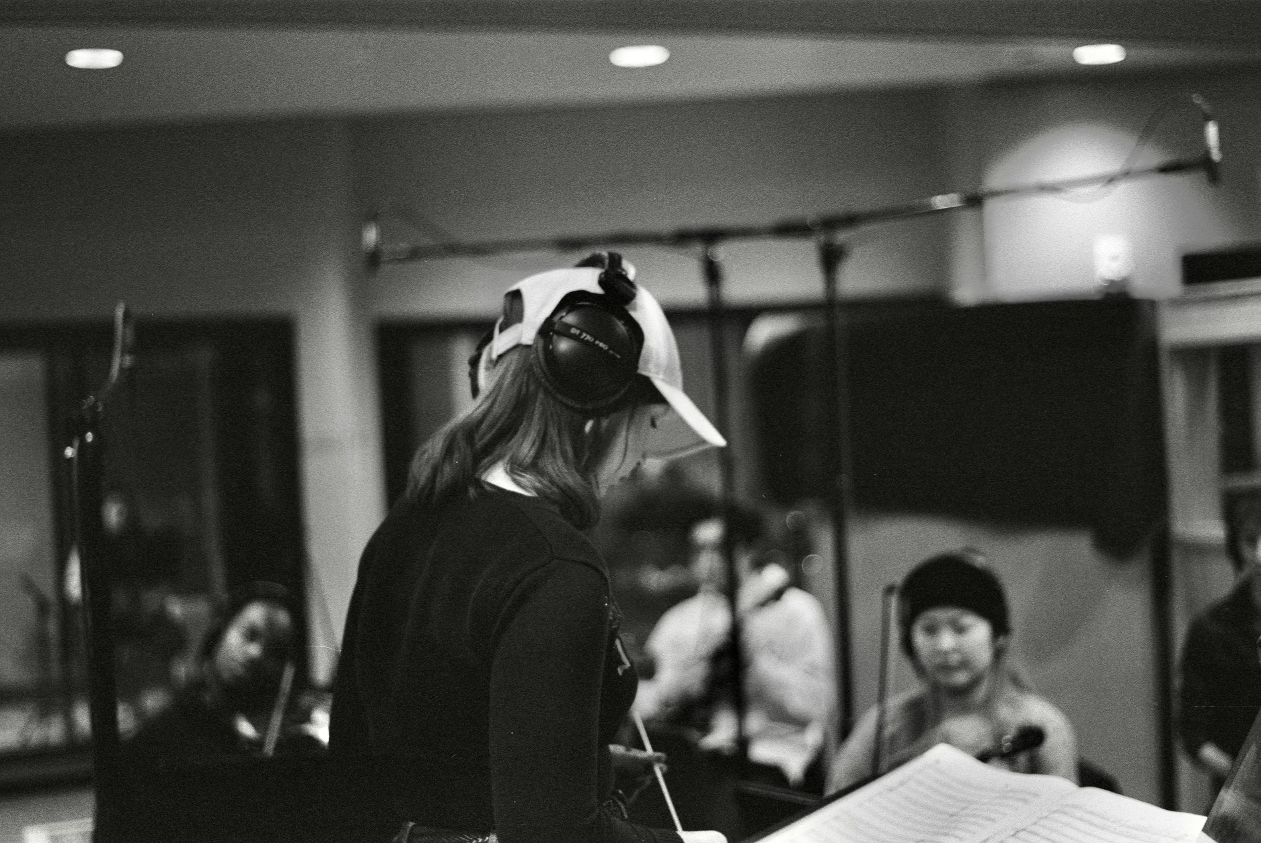 A woman with headphones, wearing a cap, standing in front of a group of people watching her read music sheets in an indoor setting.