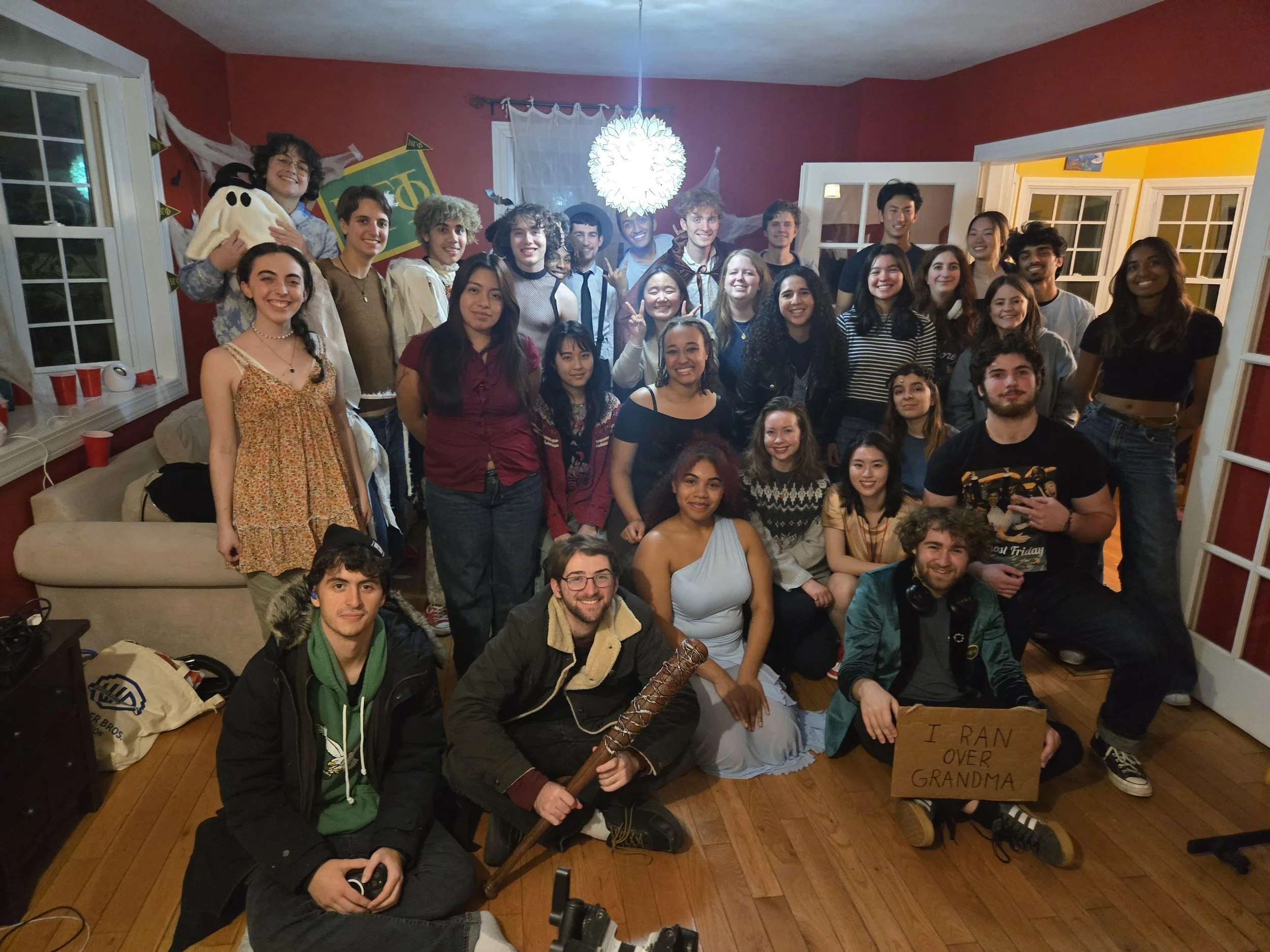 Group of people gathered indoors at a party, some dressed in costumes, with decorations and red cups visible, holding signs and props, smiling for the photo.