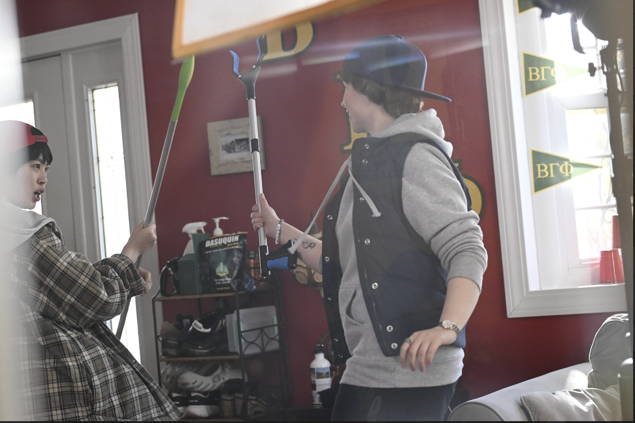Two teenagers indoors, one wearing a baseball cap backwards and a hoodie, and the other wearing a plaid jacket and a red baseball cap backwards. They are holding gardening tools and appear to be talking or preparing for an activity. There are shelves