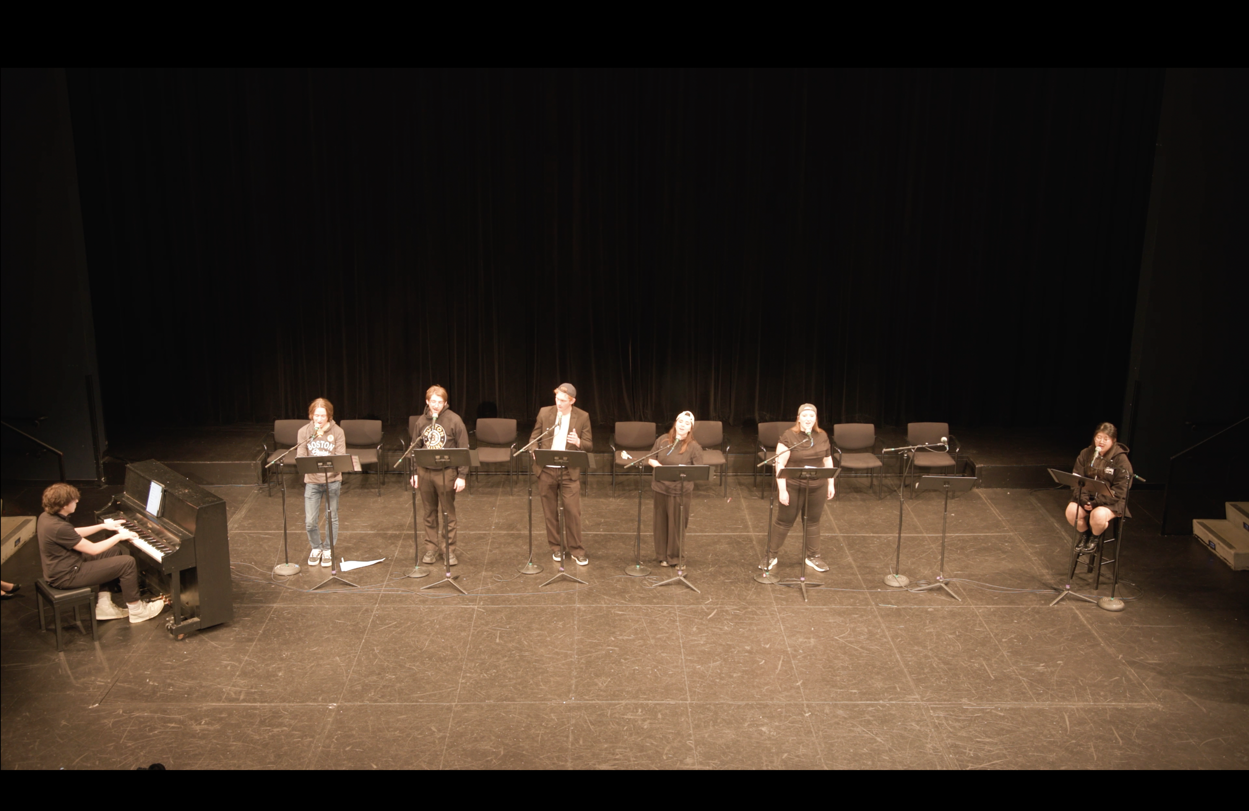 A group of performers singing for Trojan Horse's first workshop performance. They are onstage with microphones, accompanied by a pianist playing a grand piano, in a theatre with black curtains in the background.