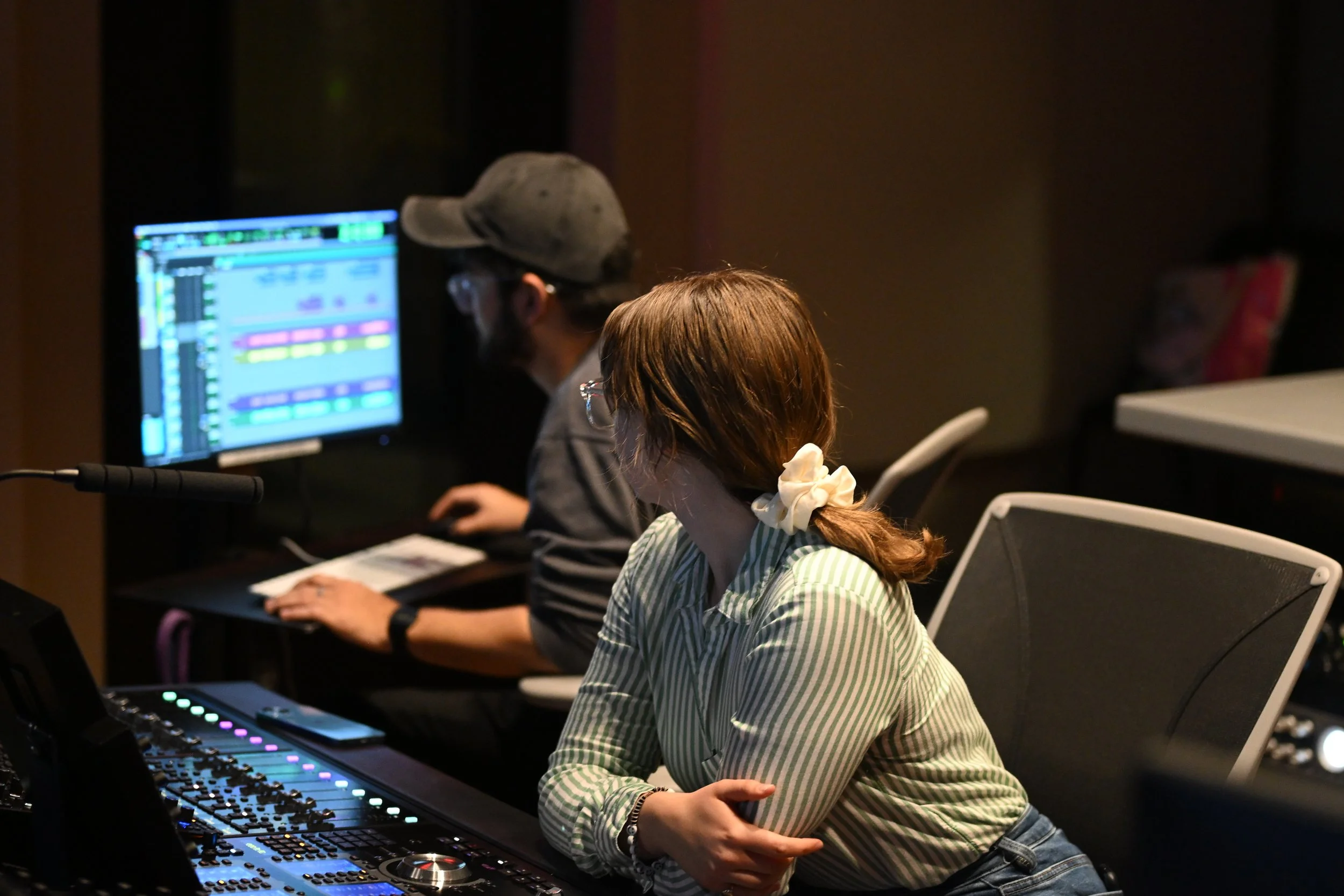 Two people working at a music production studio with a computer displaying audio editing software, a mixing console, and other electronic music equipment.