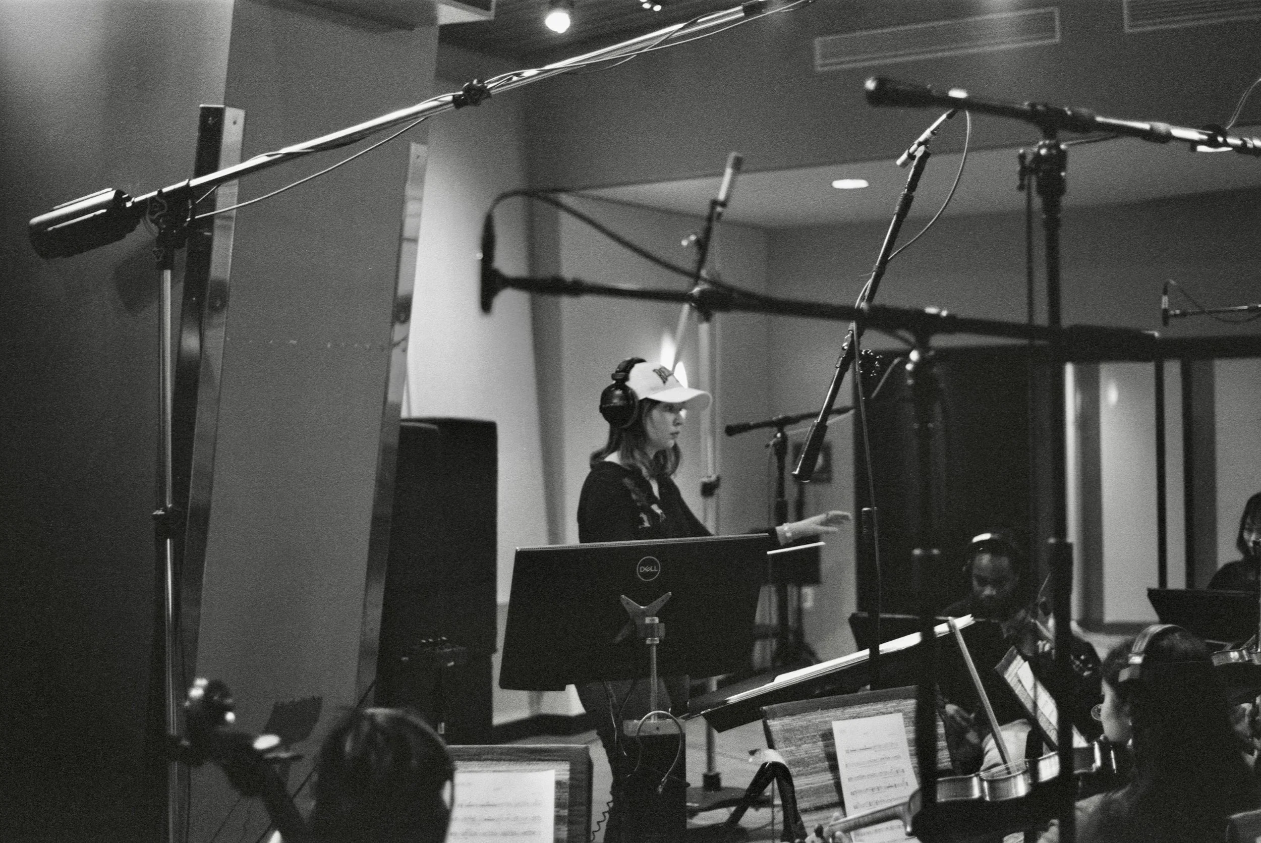 A woman wearing a hat and headphones is sitting at a desk with music sheets in front of her, surrounded by music recording and production equipment, including microphones, stands, and monitors in a recording studio.