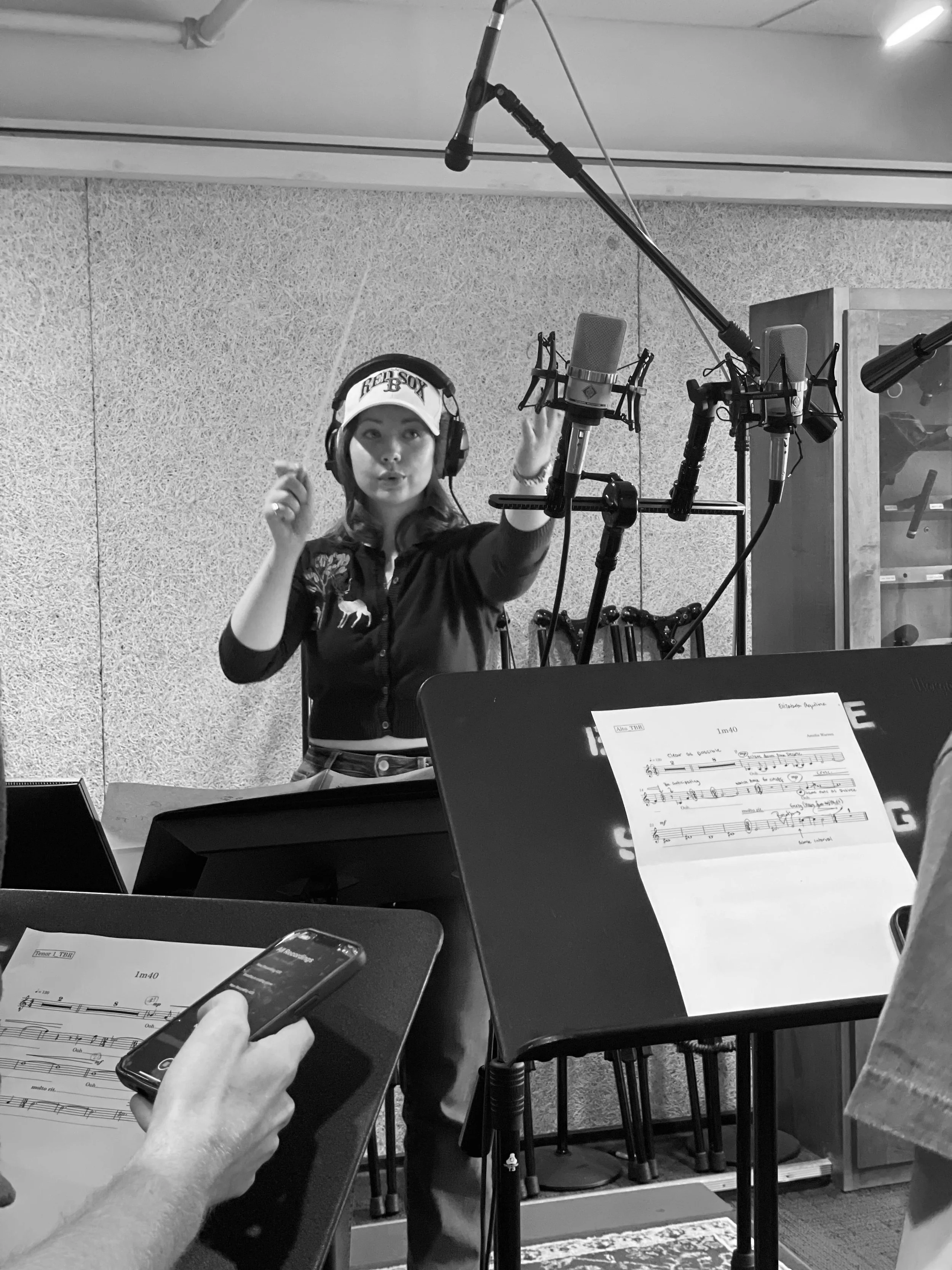 A woman in a Recording Studio, wearing headphones and a cap, stands in front of a microphone. Mixing equipment and sheet music are visible in the foreground, and a person is also holding a smartphone.
