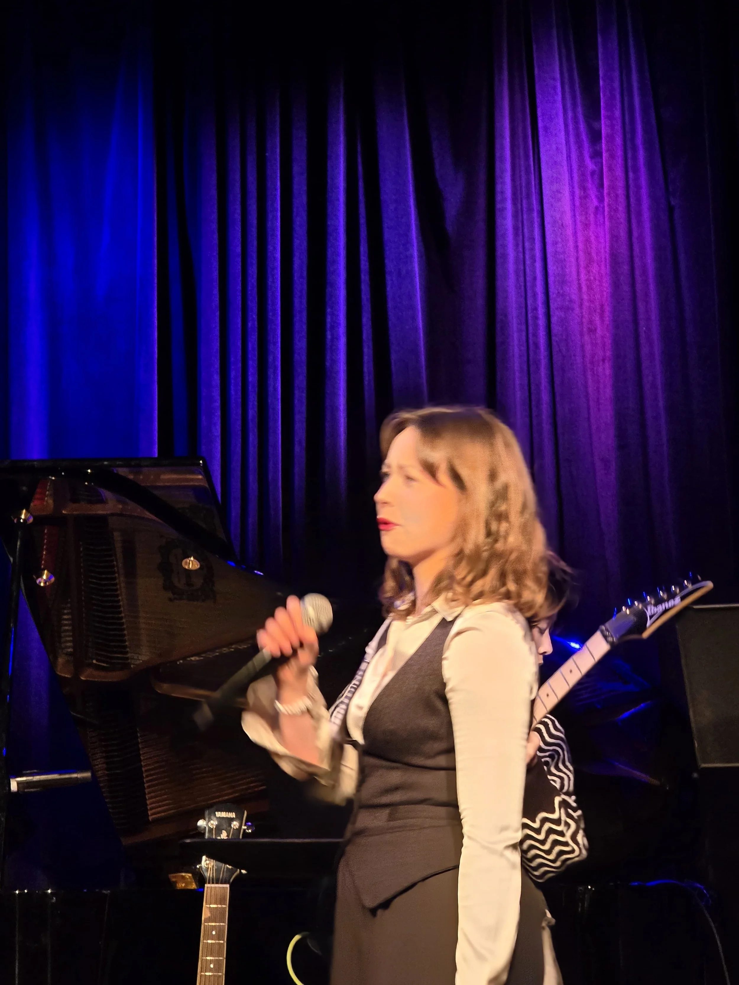 Amie Wren holding a microphone on a stage with purple curtains and a black grand piano in the background.