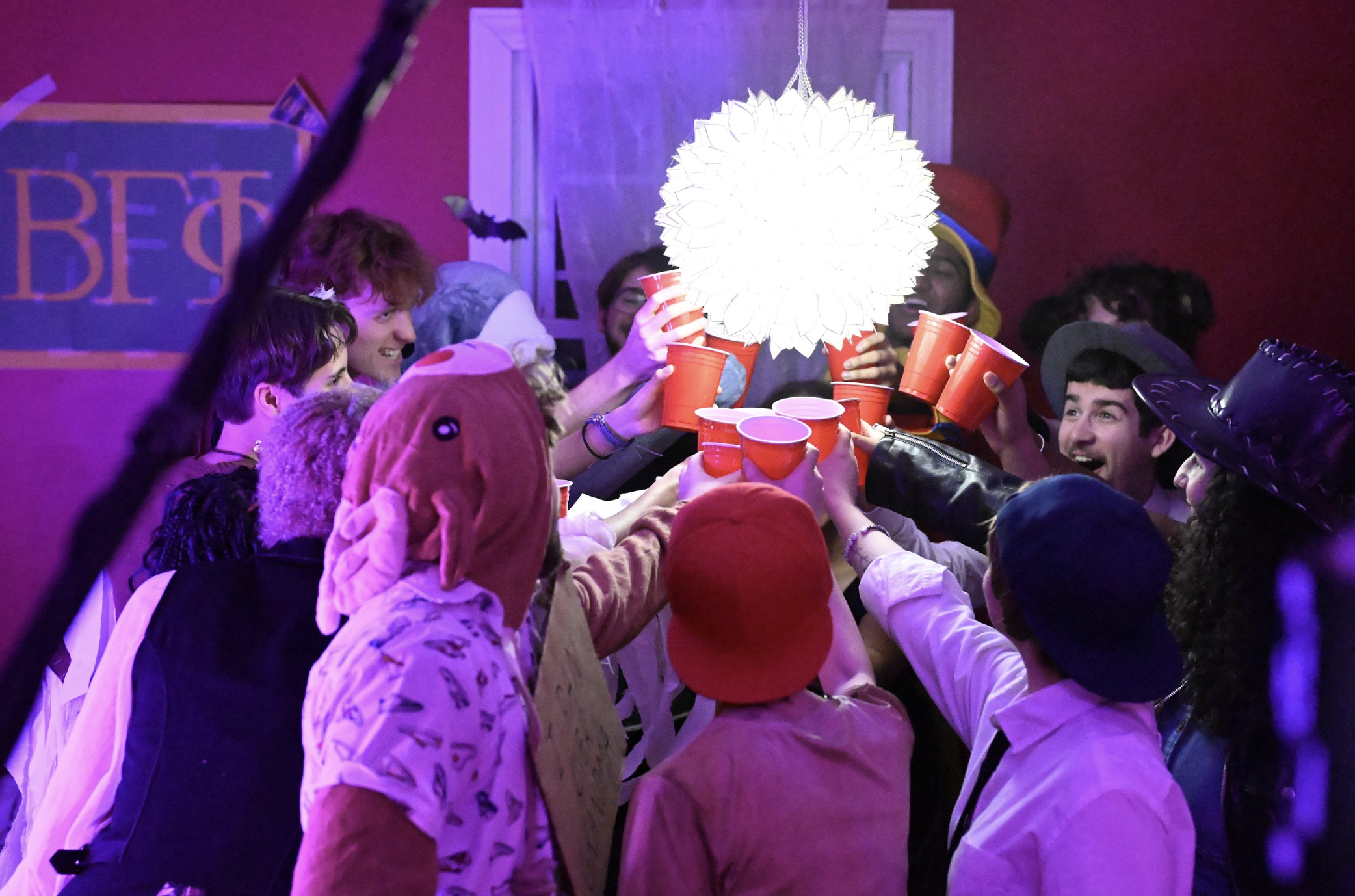A group of people dressed in costumes, raising red cups together in a celebratory toast, in a room decorated with Halloween-themed items including a large purple sign and a hanging paper lantern.