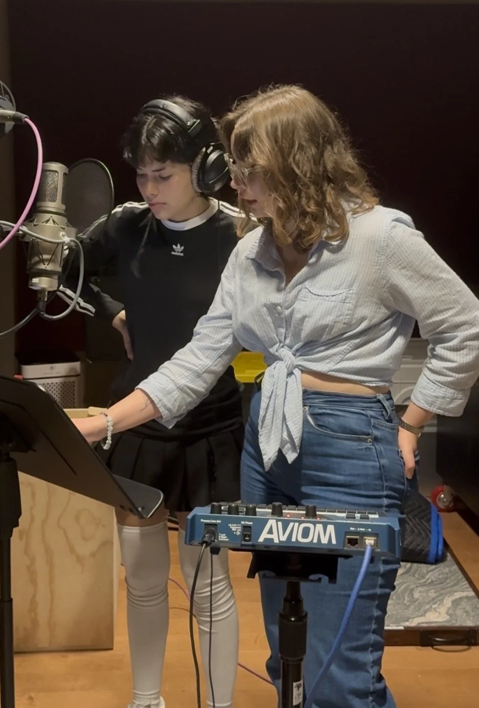 Two women working in a recording studio, one with headphones and a microphone, the other looking at a music stand and electronic equipment.
