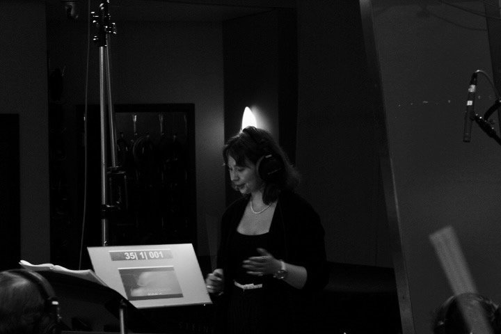 A woman in a recording studio wearing headphones, standing near a music stand and microphone, with a dark background and dim lighting.