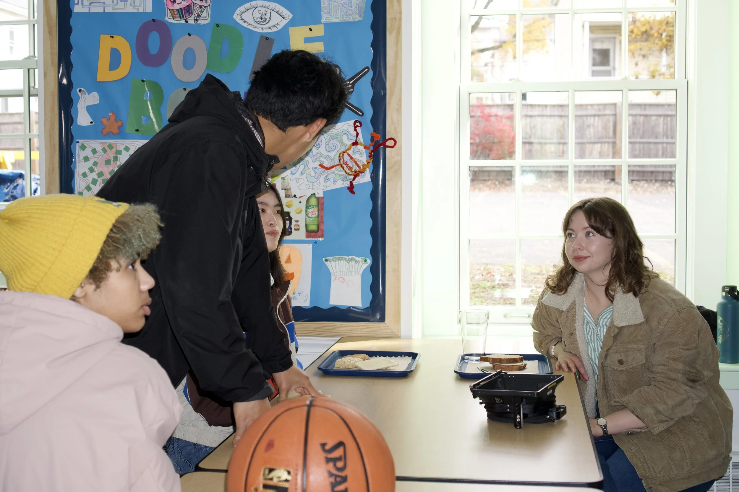 A group of young people gathered around a table with sandwiches and a basketball in a classroom, with a woman sitting on the other side of the table. The classroom has colorful posters and a window showing trees outside.