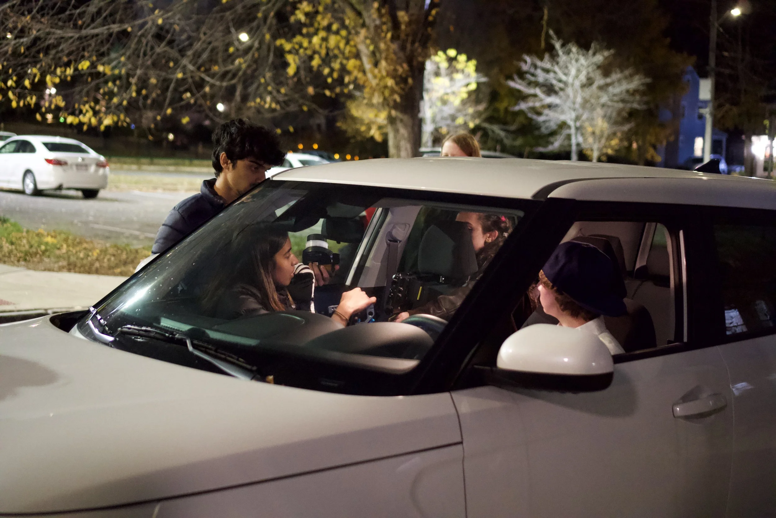 Four people gathered around the steering wheel of a white car at night, with a park and parked cars in the background, illuminated by streetlights.