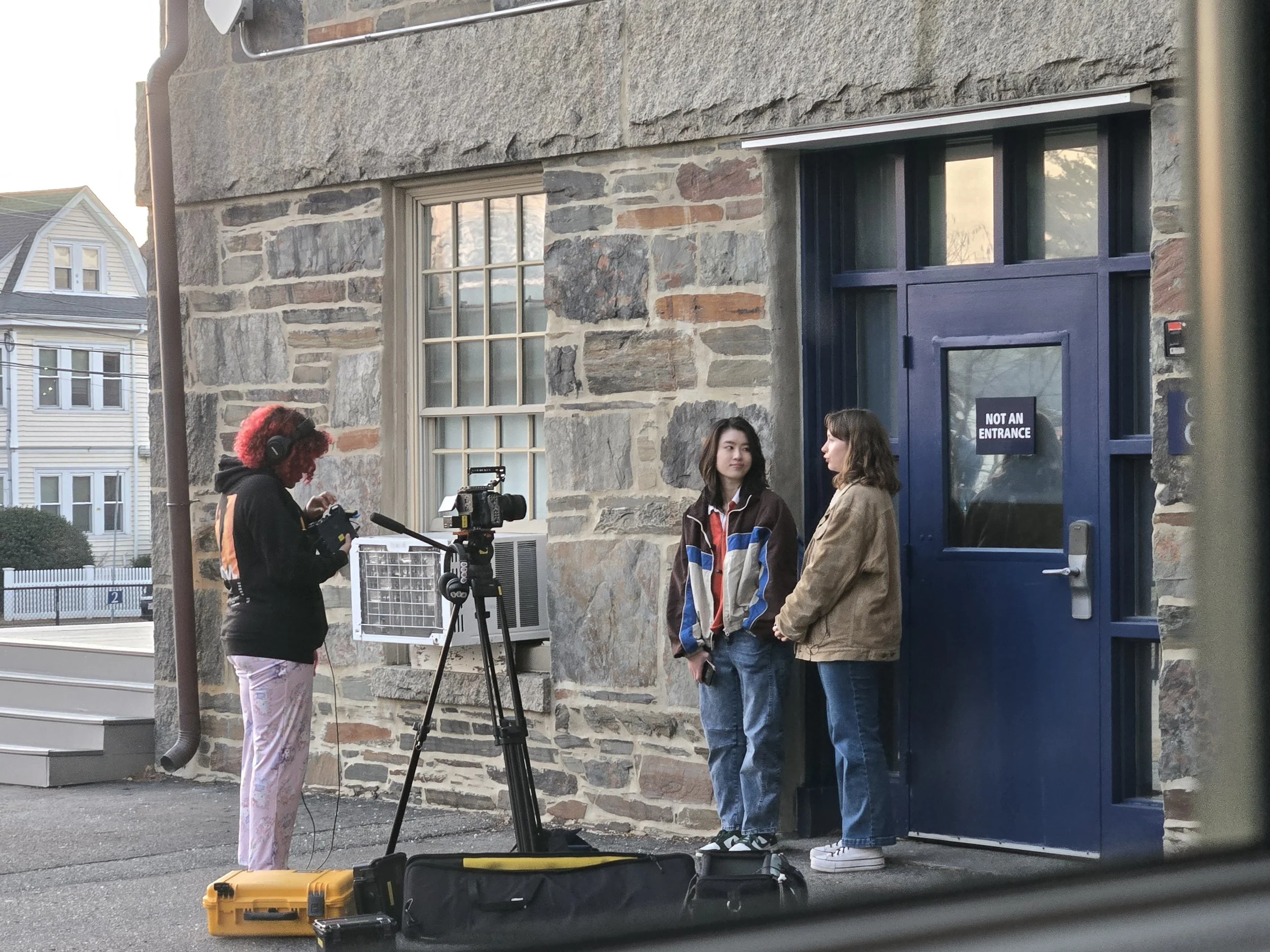 Two women and one person with pink hair, headphones and a black jacket, are preparing filming equipment outside a stone building One woman is wearing a brown jacket and jeans, and the other woman is wearing a colorful jacket and jeans. A camera on a 