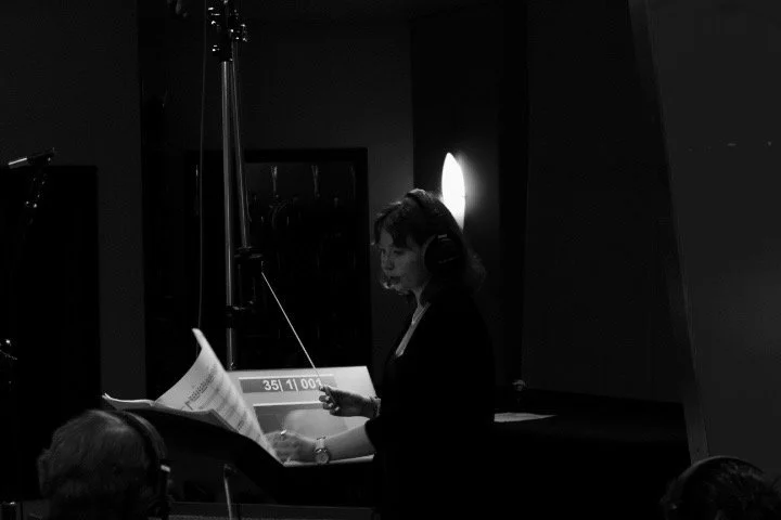 A woman in a recording studio wearing headphones, reading sheet music or a script, with a microphone and soundproofing panels around her.