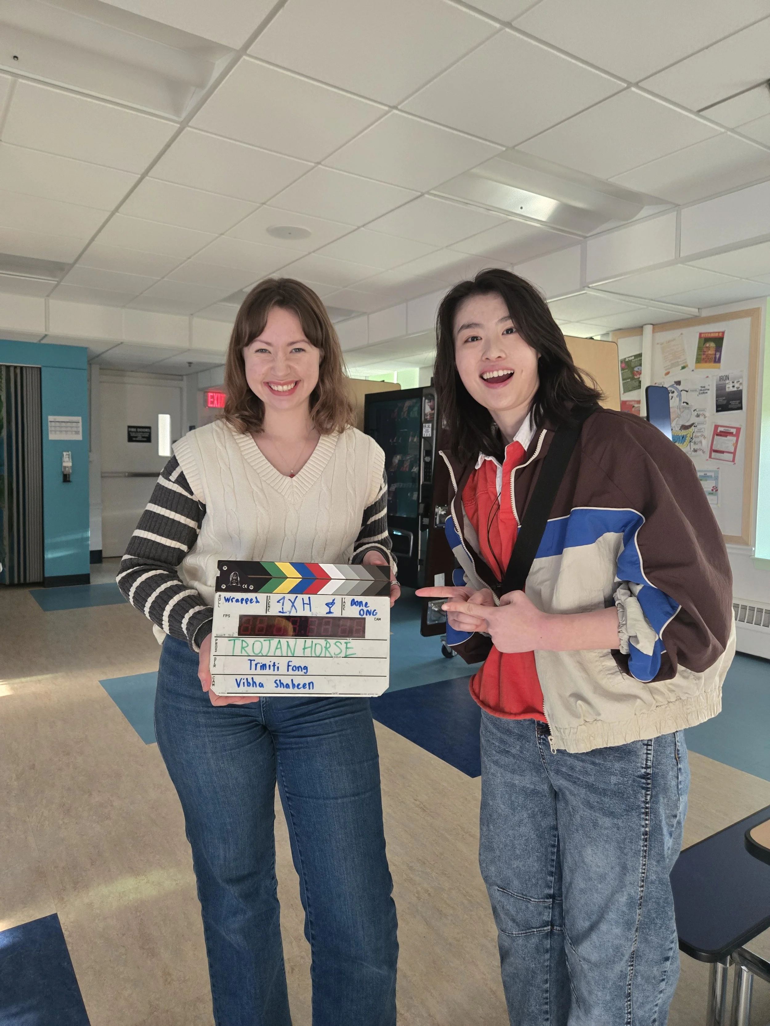 Two women standing indoors, smiling, with one woman holding a film slate. The woman on the left has shoulder-length brown hair, wearing a cream sweater with black and white striped sleeves, and blue jeans. The woman on the right has medium-length bla