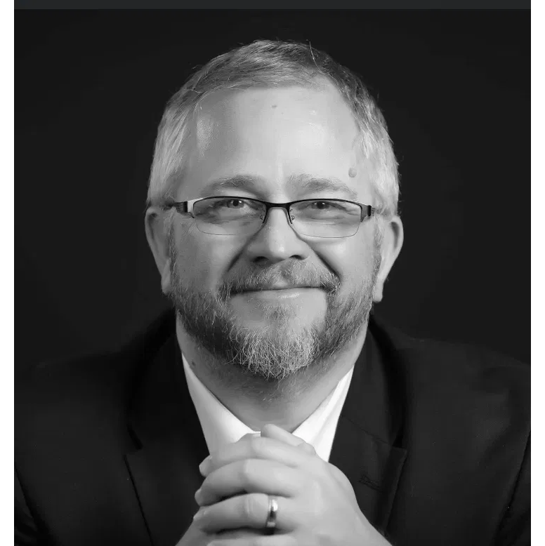 A black-and-white portrait of a man with glasses, a beard, and short hair, smiling and wearing a suit jacket and white shirt, with hands clasped in front of him