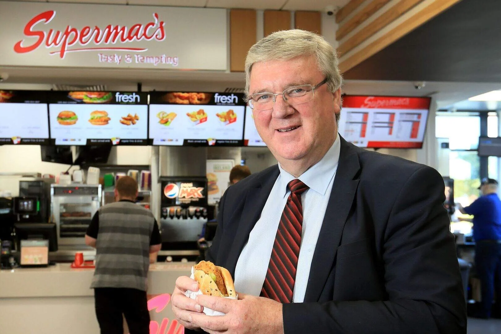 A man in a suit and tie holding a fast food sandwich inside a fast food restaurant with a "Supermac's" sign and digital menu screens in the background.