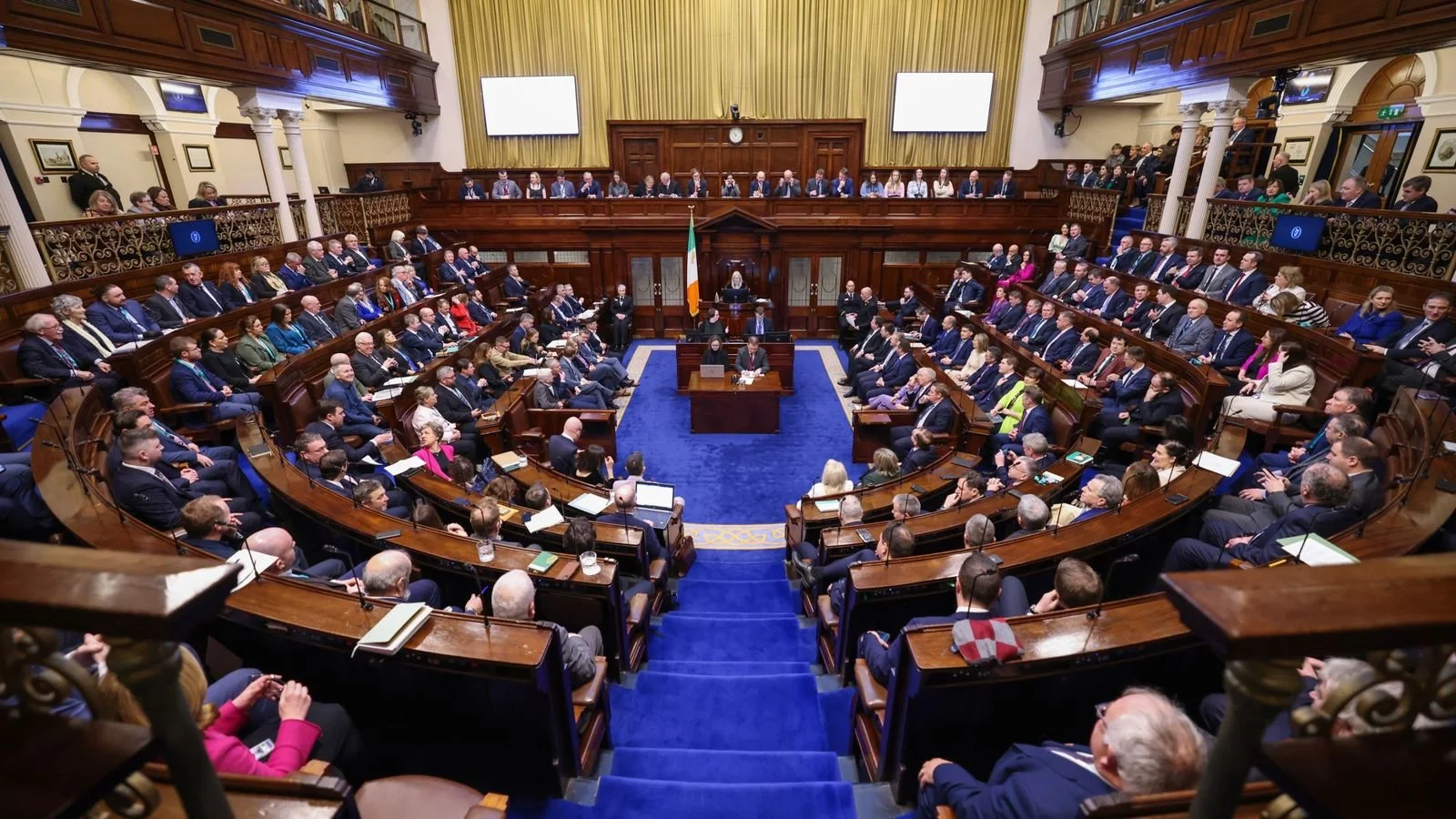 A large parliamentary chamber filled with many people seated in a semi-circular arrangement, with a central speaker at a podium in front of an Irish flag, and two large screens on the walls behind.