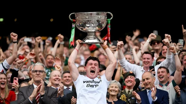 A soccer player holding a large trophy above his head, celebrating with a crowd and team members in the background, some taking photos and cheering.