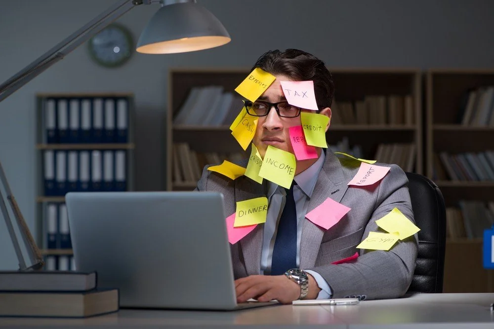 A man in a suit and glasses sitting at a desk covered with numerous sticky notes with words like 'expenes,' 'tax,' 'income,' and 'dinner,' looking frustrated while working on a laptop.
